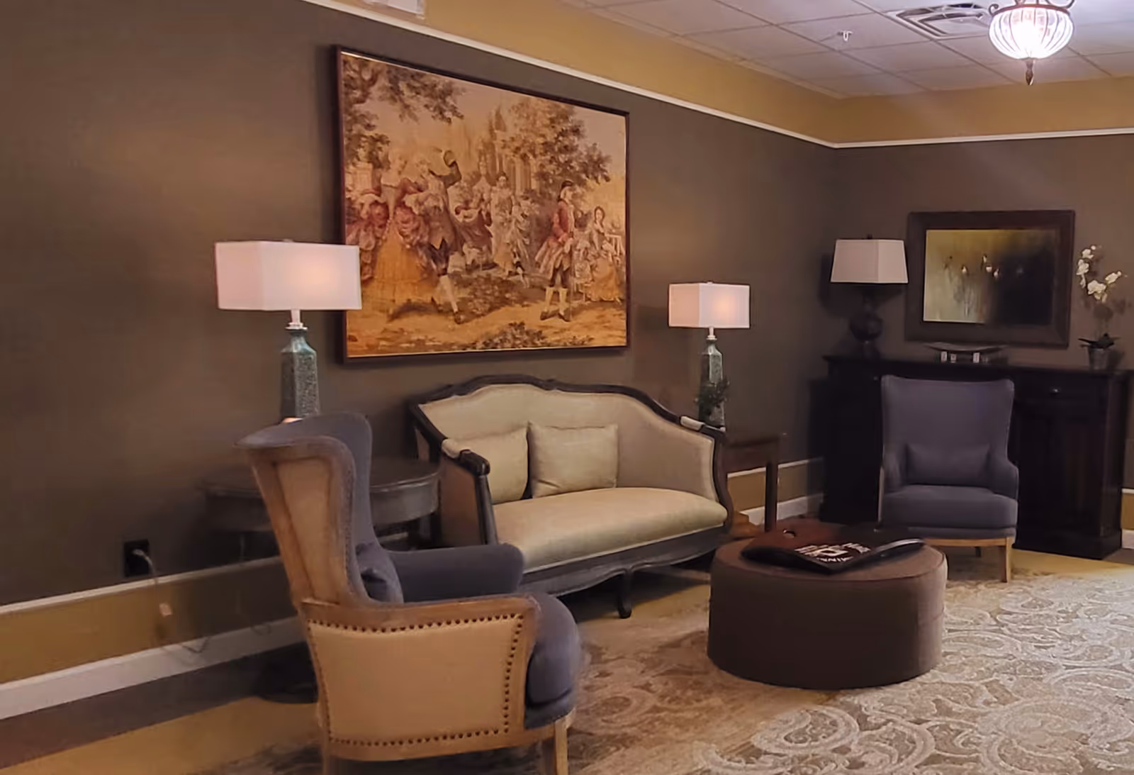 A cozy living room area with a beige loveseat, two gray armchairs, a round ottoman with a magazine on top, two table lamps on side tables, a dark wooden cabinet with a lamp and decorative items, and two framed artworks on the walls. The room has warm brown walls and a patterned rug on the floor.