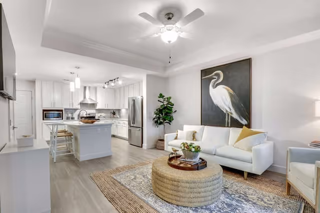 A bright and modern open-concept living room and kitchen area. The living room features a white sofa with yellow and white pillows, a round woven coffee table with decorative items, a large framed painting of a white heron on the wall, and a potted plant in the corner. The kitchen has white cabinetry, stainless steel appliances including a refrigerator and range hood, a kitchen island with bar stools, and pendant lighting. The flooring is light wood, and the ceiling has a white fan with a light fixture.