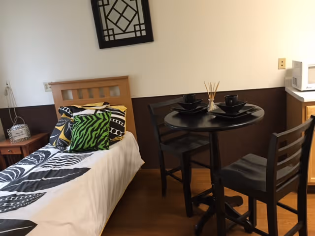 A small supportive living room featuring a single bed with patterned pillows and a black and white bedspread, a round black table set with black dishes and two chairs, a small wooden nightstand with a decorative lantern, and a microwave on a wooden cabinet against the wall.