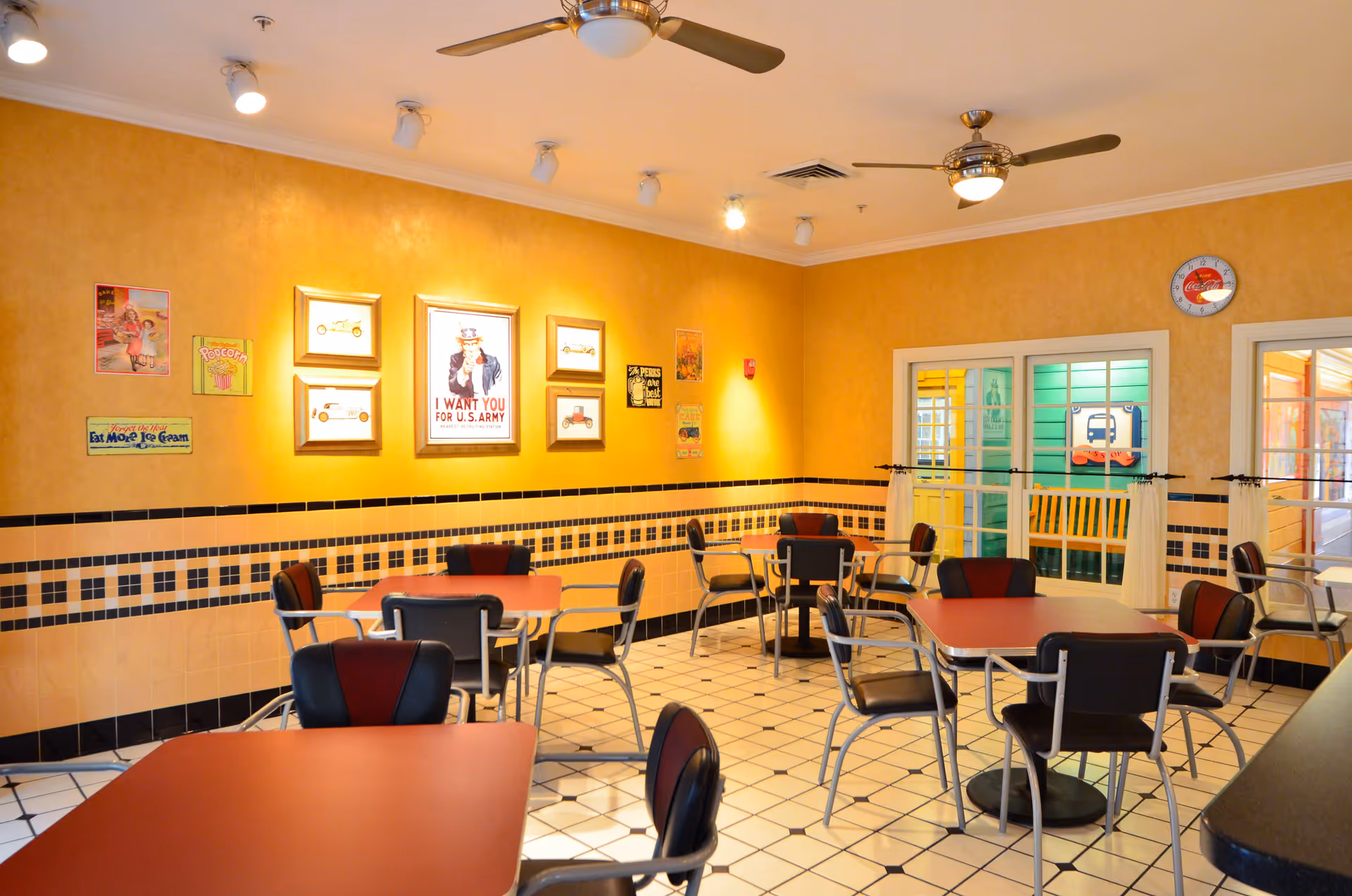 Bright dining room with red-topped tables, black chairs, tiled floor, yellow walls decorated with framed posters, and ceiling fans.