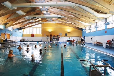 Indoor swimming pool with several people participating in a water exercise class. The pool area has a high wooden beam ceiling with large windows letting in natural light. There are poolside benches and equipment visible along the sides.