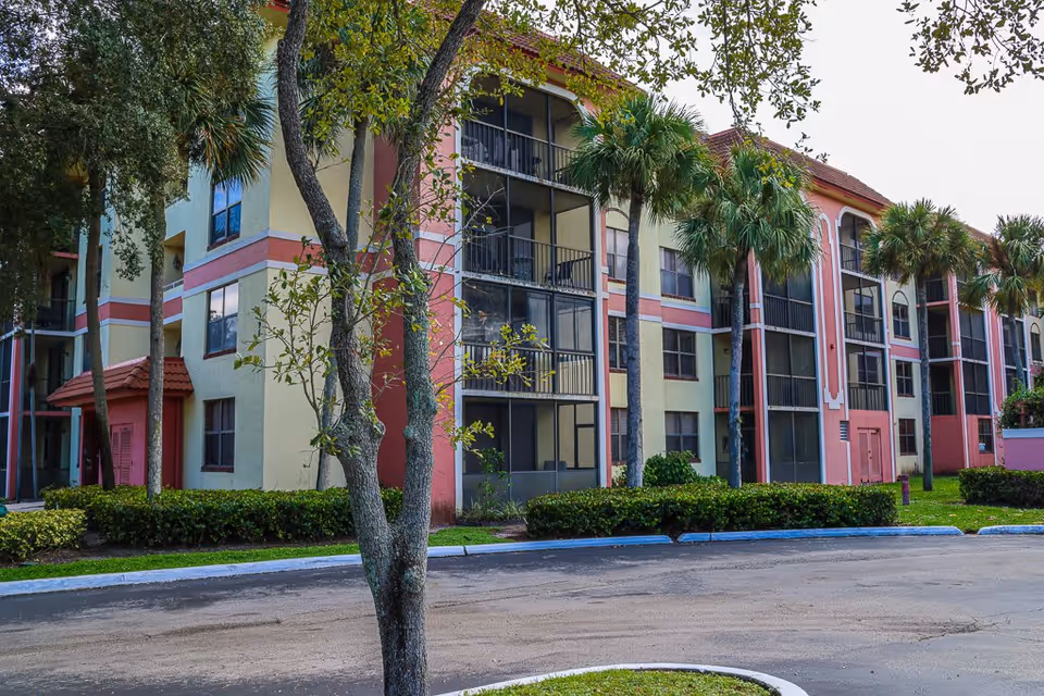 Exterior view of a multi-story residential building with balconies, surrounded by palm trees and greenery, under a clear sky.