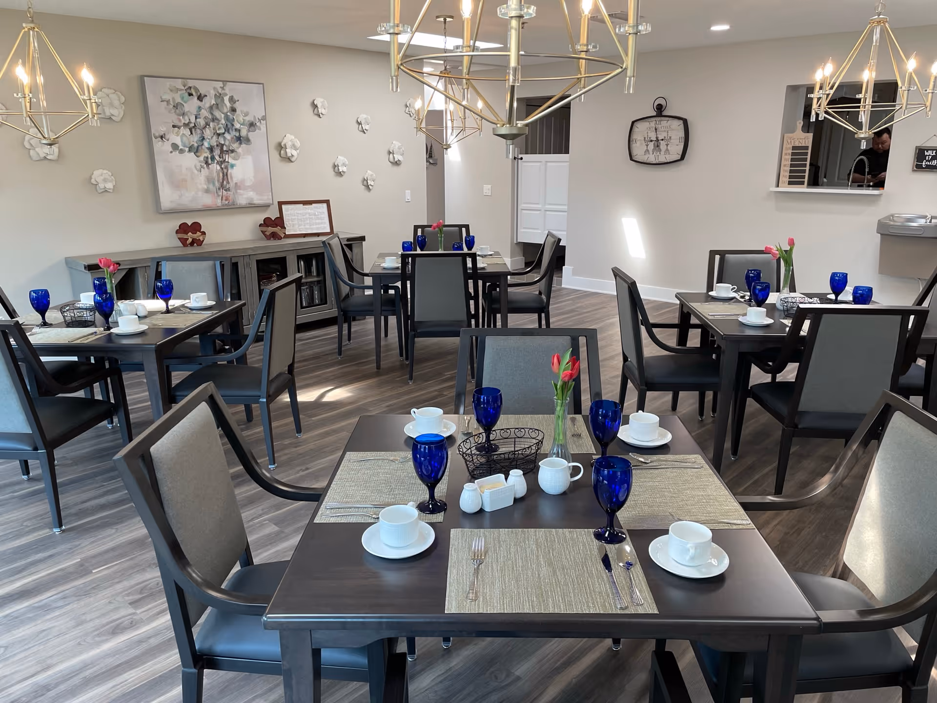 A bright dining room with several dark wood tables set with blue water glasses, white cups and saucers, and small vases holding pink tulips. The room has light-colored walls decorated with white flower wall art and a large abstract painting. Modern chandeliers hang from the ceiling, and there is a sideboard against the wall with decorative items. The floor is covered with wood-style flooring.