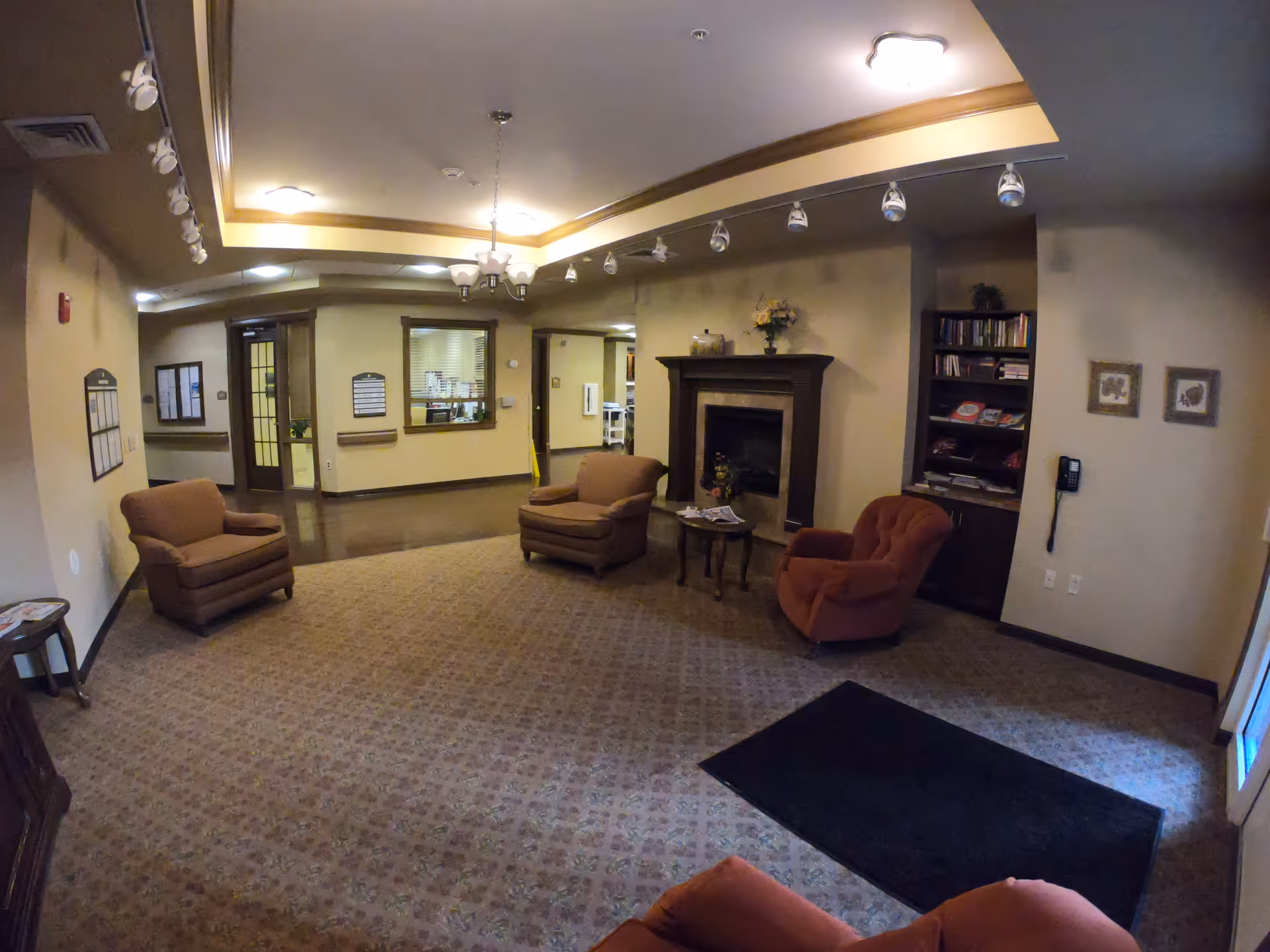 A cozy lobby/lounge with armchairs arranged around a fireplace, a bookshelf, and a reception window in the background.