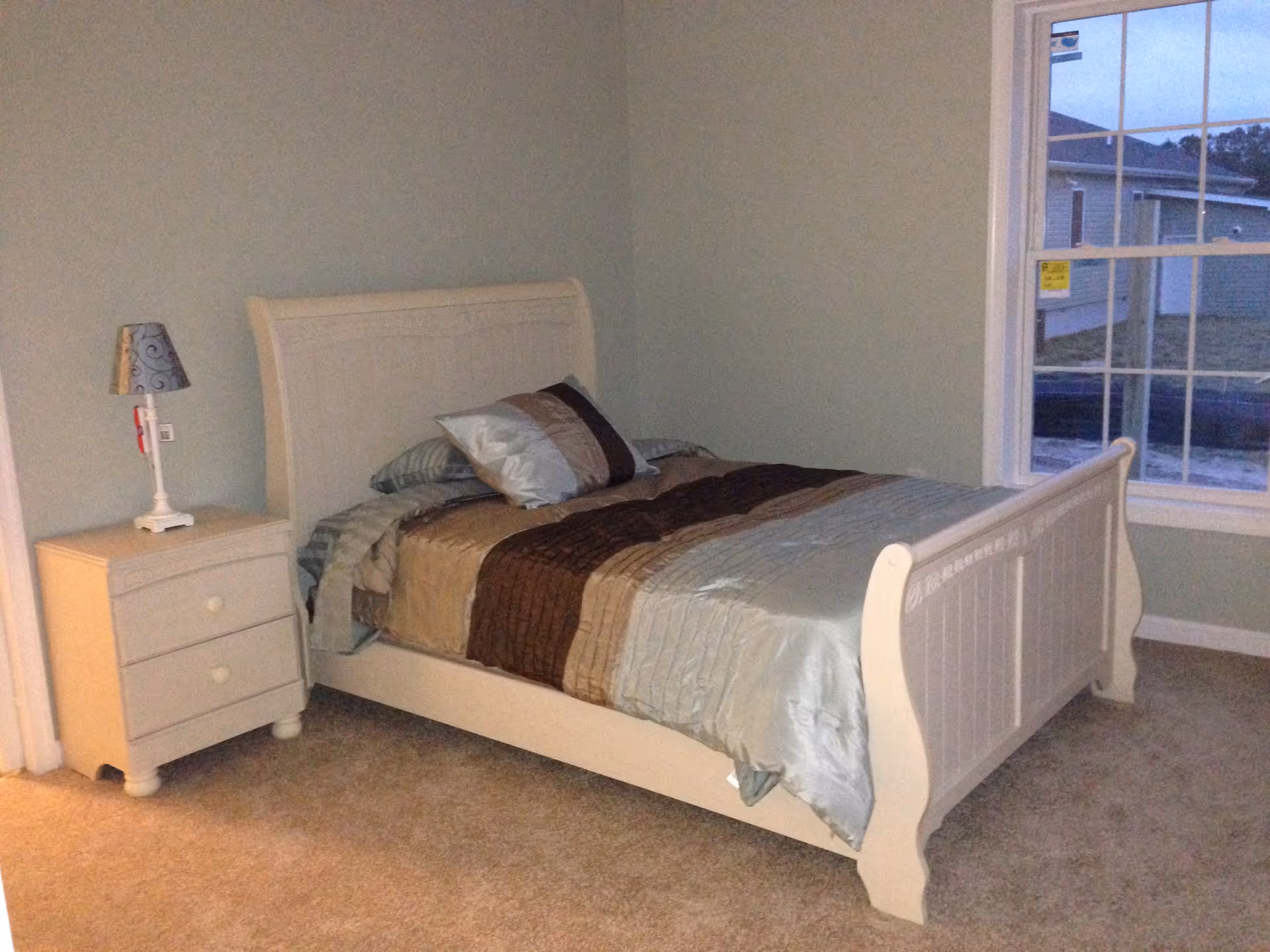 A simple bedroom with a white wooden bed made with brown and gray bedding, a matching nightstand and lamp, carpeted floor, and a window looking outside.