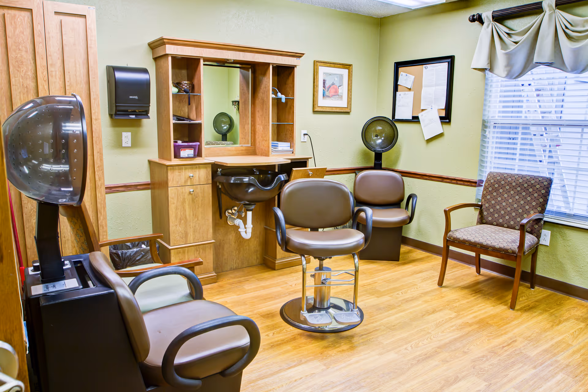 Interior of a hair salon room in a senior living facility with a hair dryer chair, a styling chair in front of a wooden vanity with a mirror and sink, two additional chairs, a bulletin board with papers, and a window with blinds and draped curtains.