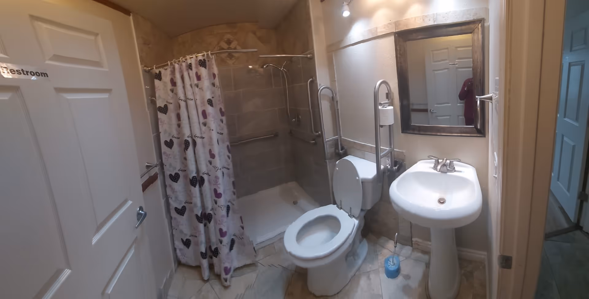 A small bathroom with a white toilet, a pedestal sink, and a walk-in shower with a curtain decorated with hearts. The shower area has tiled walls and grab bars for accessibility. A mirror is mounted above the sink, and the door to the bathroom is partially open with a sign that reads 'Restroom'.
