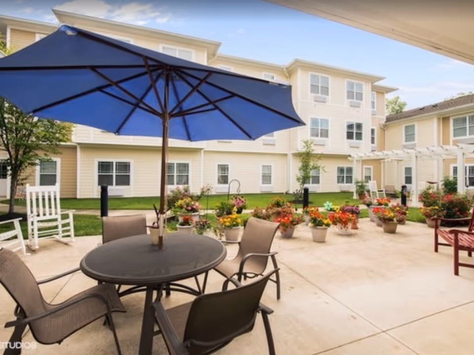 Courtyard patio with a round table, chairs and a large blue umbrella surrounded by potted flowers in front of a multi-story residential building.