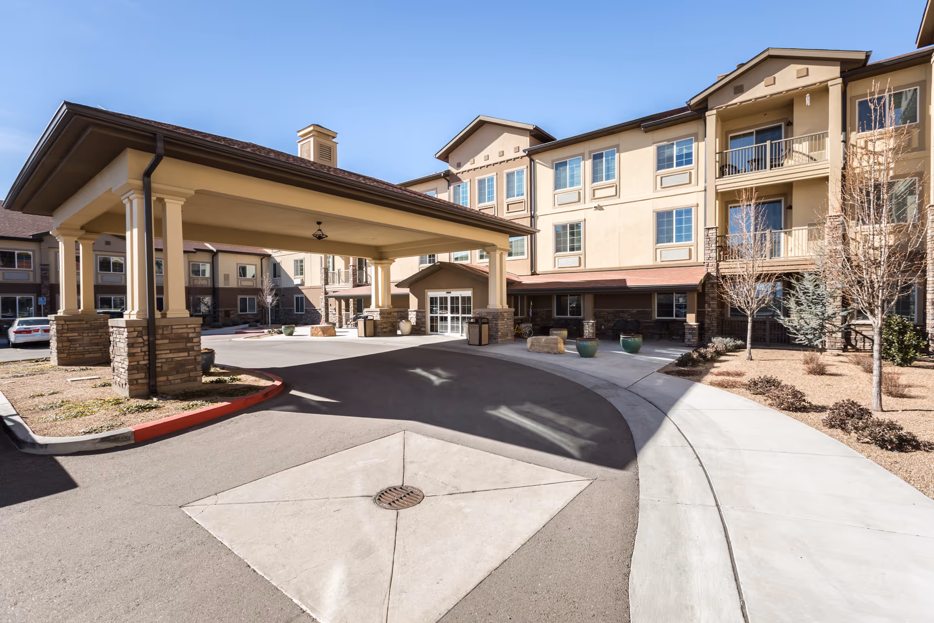 Exterior view of Palmilla Senior Living facility showing a covered driveway entrance with stone pillars, a multi-story beige building with balconies and windows, and a landscaped area with small trees and shrubs under a clear blue sky.