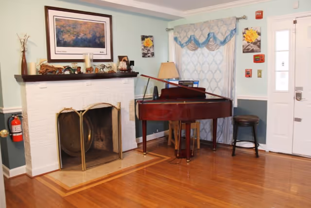 Cozy common room with a white brick fireplace, mantel decor, a grand piano, hardwood floors, and an entry door.