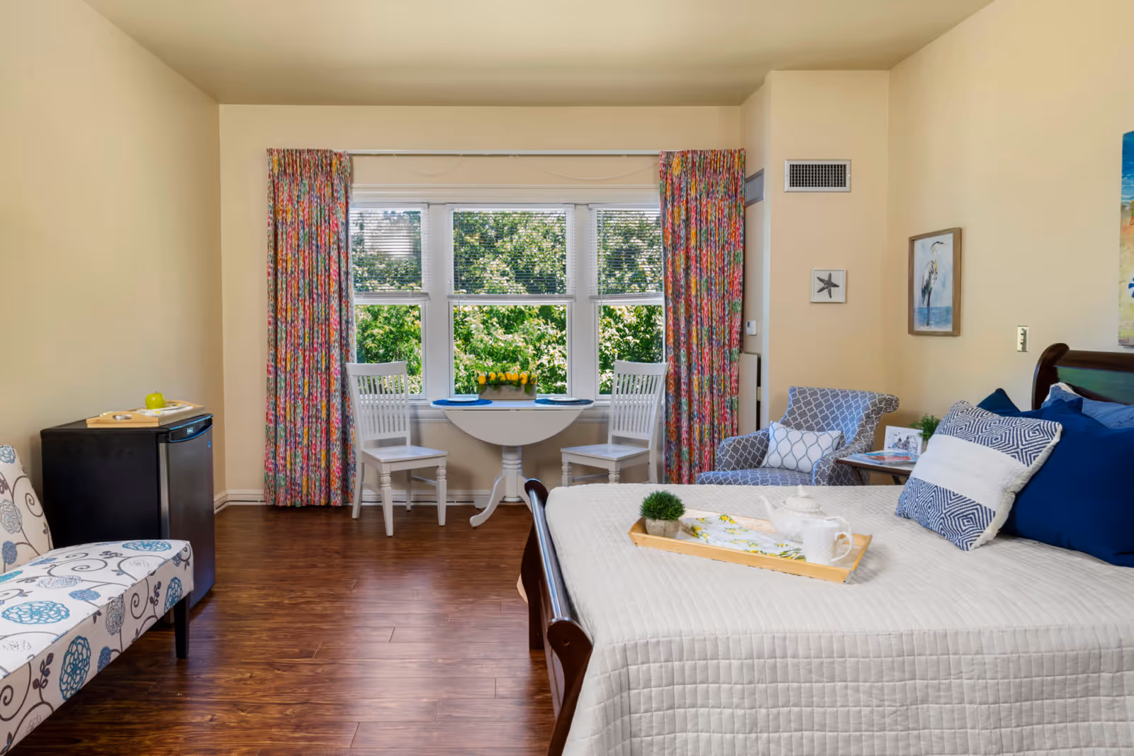 A bright and cozy bedroom with a large bed covered in a white quilt and multiple blue and white pillows. There is a wooden tray with a teapot, cup, and small plant on the bed. To the right of the bed is a patterned armchair with a pillow and a small side table with a photo frame and plant. In front of a large window with colorful floral curtains, there is a small white round table with two white chairs. On the left side of the room, there is a patterned bench and a small black mini-fridge with a tray on top. The room has wooden flooring and light beige walls with framed artwork.