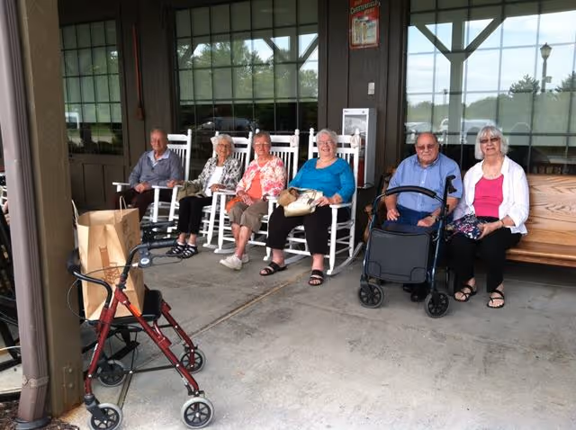 Six elderly people sitting on rocking chairs and a wooden bench on a covered outdoor patio area. Two walkers are visible in the foreground. The background shows large windows and a wooden structure.