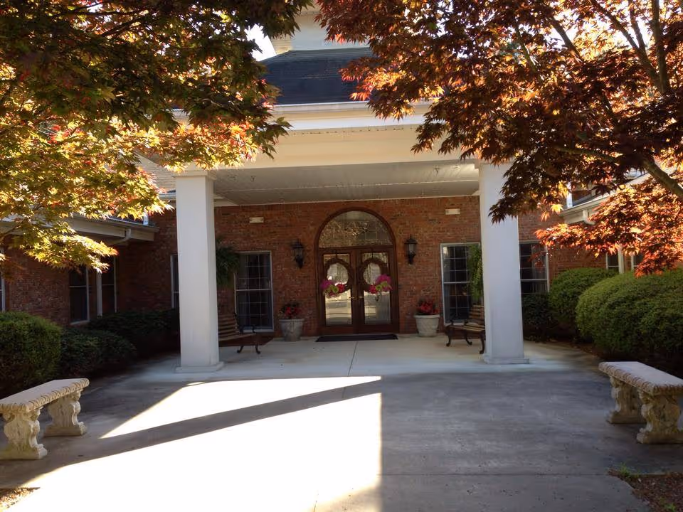 Entrance of Southern Estates Assisted Living Community showing a covered porch with two white columns, a double wooden door with glass panels decorated with wreaths, two benches on either side, potted plants, and trees with autumn-colored leaves framing the scene.