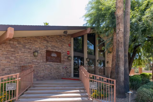Entrance to Heritage Court Post Acute Of Scottsdale featuring a stone facade, large glass windows, a ramp with railings on both sides, and surrounding greenery including trees and bushes.