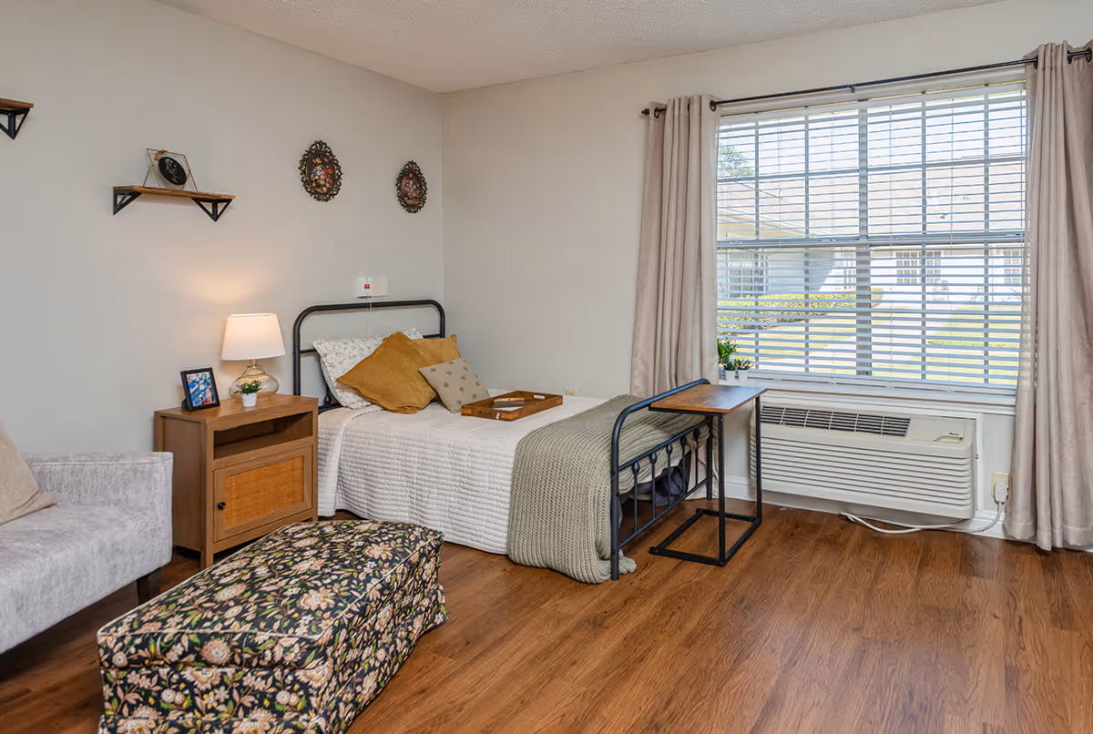 A cozy bedroom with a single bed featuring white bedding and multiple pillows, a wooden nightstand with a lamp and small plant, a floral patterned ottoman, a beige armchair, and a large window with beige curtains letting in natural light. The room has wooden flooring and light-colored walls with decorative plates hanging above the bed.