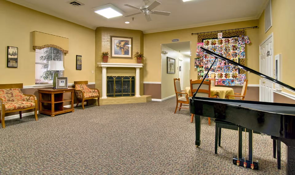 A cozy common area in a senior living facility featuring a black grand piano in the foreground, a round table with four chairs covered with a yellow tablecloth, a colorful quilt hanging on the wall, a fireplace with a painting and two potted plants on the mantel, two floral upholstered chairs, a wooden side table with a lamp and framed photo, and beige walls with carpeted flooring.