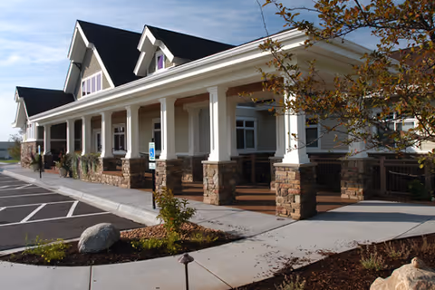 Front exterior of an assisted living building with a covered porch supported by columns and stone pillars, parking spaces, and landscaping.
