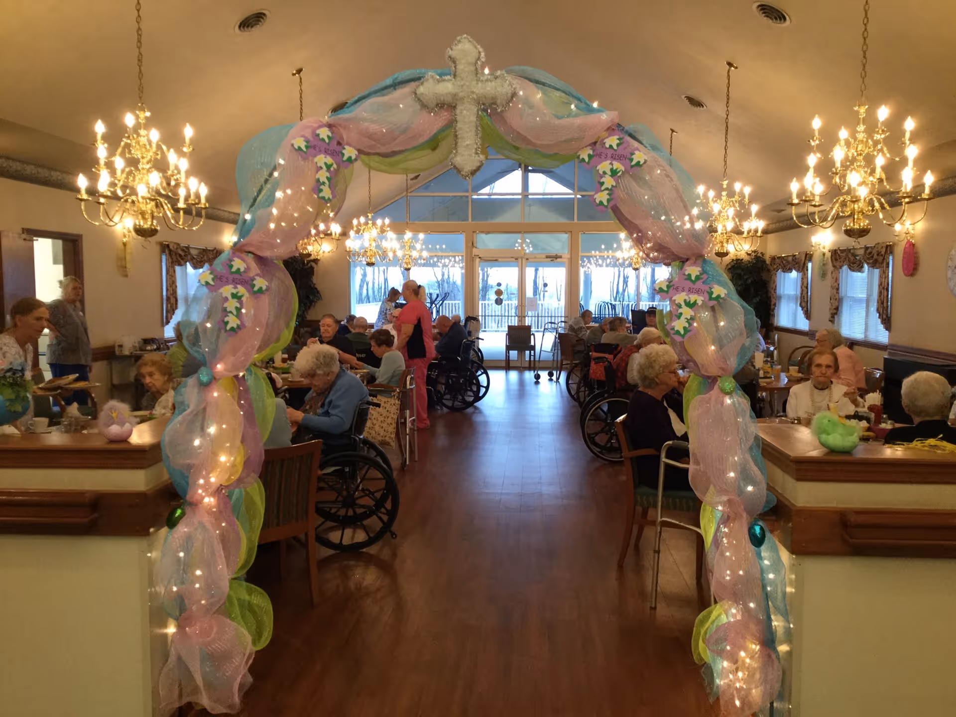 Communal dining room with a pastel-decorated archway and residents seated at tables and in wheelchairs.