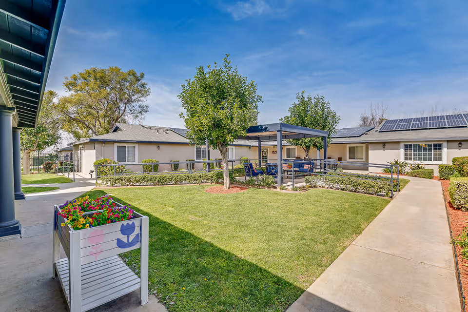 Outdoor courtyard area at Cottages at Riverside featuring a green lawn, two small trees, a covered seating area with chairs and tables, a flower planter with colorful flowers, and surrounding single-story buildings with solar panels on the roofs under a clear blue sky.