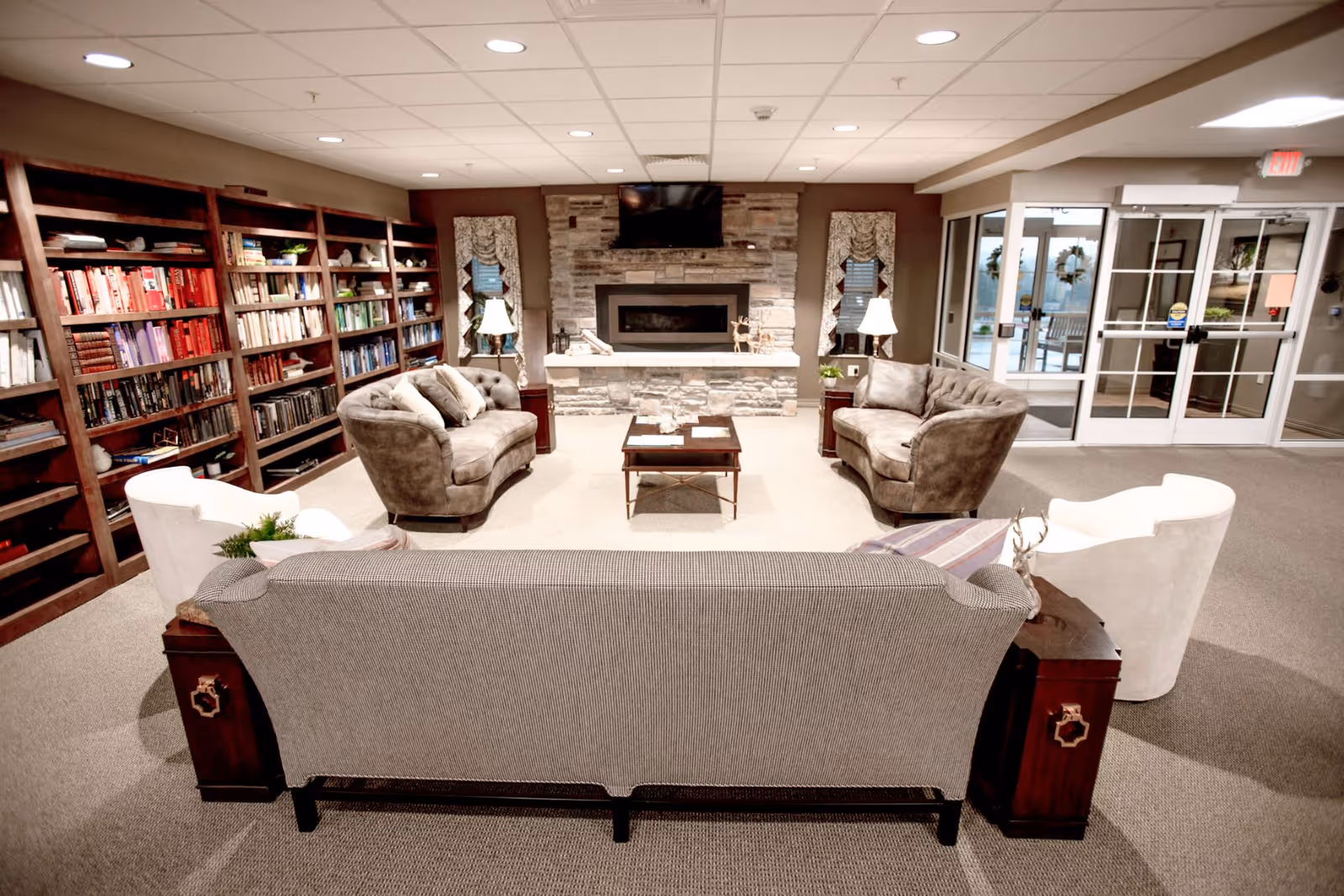 A cozy communal lounge with sofas and armchairs arranged around a coffee table and stone fireplace, with bookshelves along one wall and an entrance door visible.