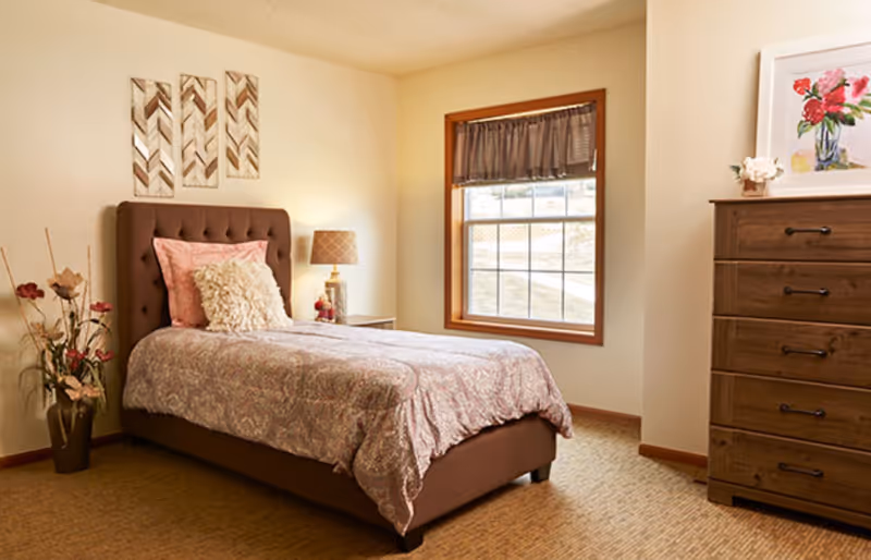 A cozy bedroom with a single bed featuring a tufted brown headboard, pink and cream pillows, and a patterned bedspread. Next to the bed is a small nightstand with a lamp and decorative items. A window with a brown valance lets in natural light. On the right side, there is a wooden dresser with a framed floral painting and a small flower arrangement on top. A vase with artificial flowers is placed on the floor beside the bed. The walls are light-colored and the floor is carpeted.
