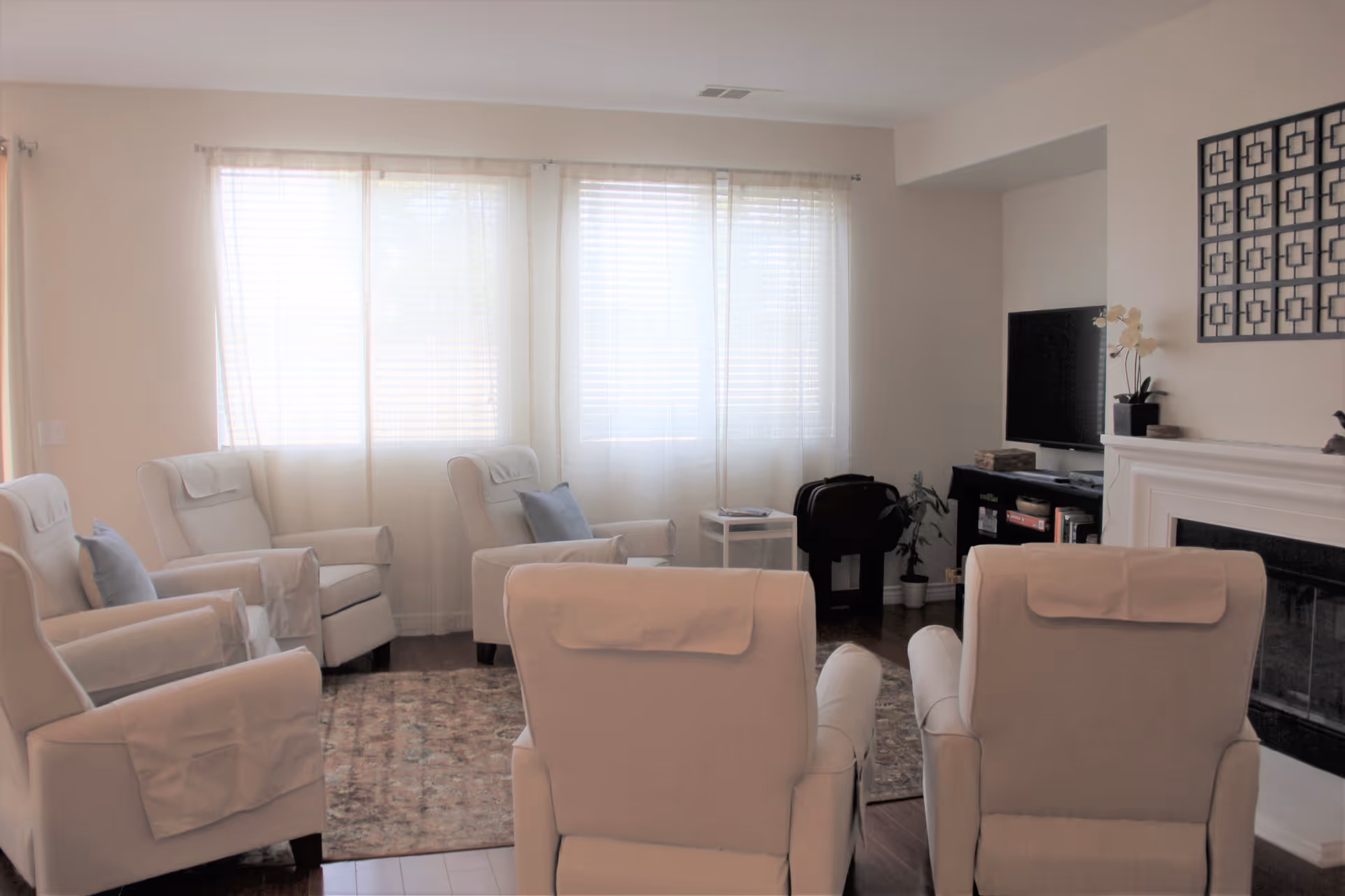 Bright living room with several upholstered recliners arranged around a rug facing a TV and fireplace under sheer curtains.