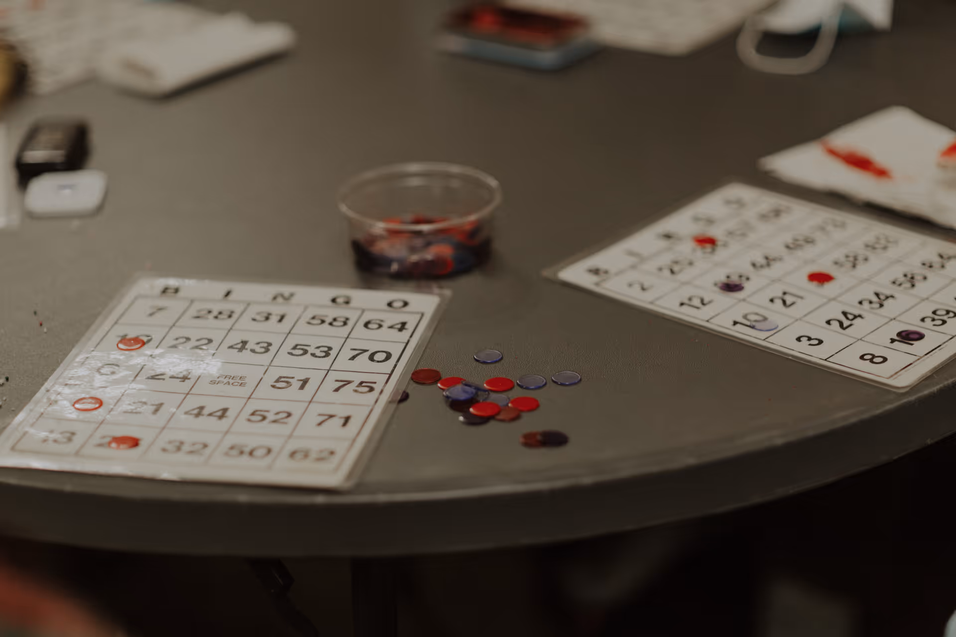 Close-up view of a table with bingo cards, red and purple bingo chips, and a small plastic container holding additional chips.