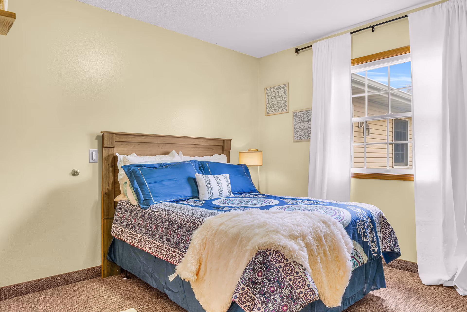 A cozy bedroom with a wooden headboard bed dressed in blue and patterned bedding, a fluffy cream-colored throw blanket, and multiple pillows. The room has beige walls, a window with white curtains, and two decorative wall hangings. A lamp is visible in the corner near the window.
