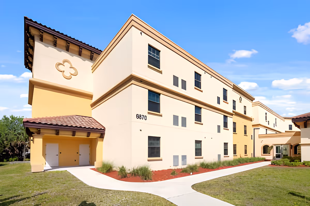 Exterior view of a three-story beige and yellow building with multiple windows, a tiled roof, and a pathway leading to the entrance. The sky is clear with a few clouds, and there is grass and landscaping around the building.