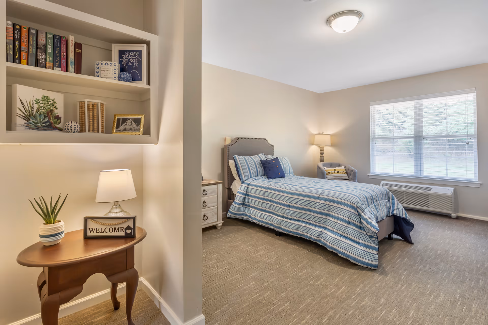 Well-lit furnished bedroom with a twin bed dressed in striped bedding, a nightstand, window, and a small table with a lamp and 'WELCOME' sign.
