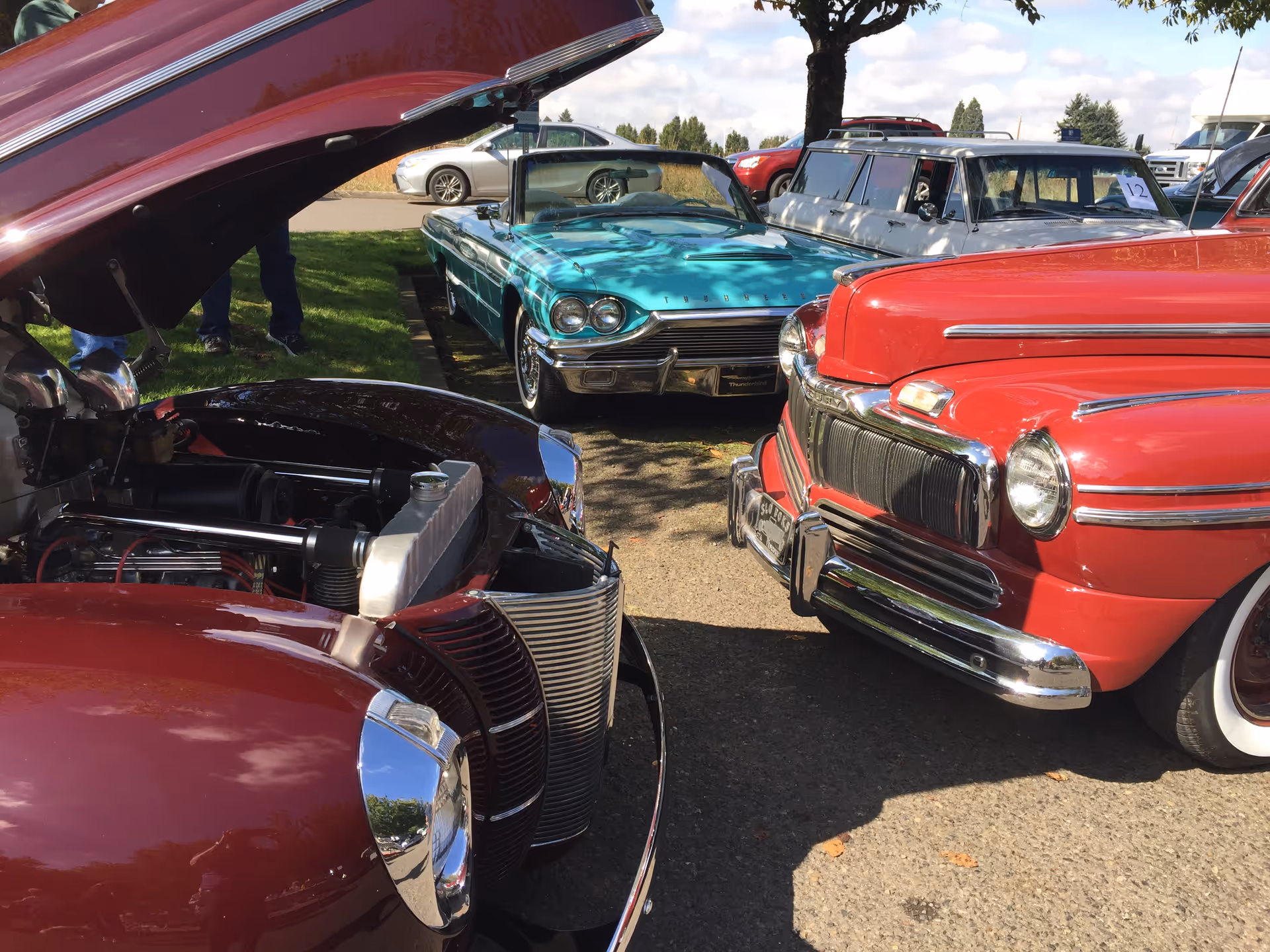 A collection of vintage cars parked outdoors on a sunny day. The image shows a maroon car with its hood open in the foreground, a turquoise convertible car in the middle, and a red classic car on the right. Other cars and a tree are visible in the background.
