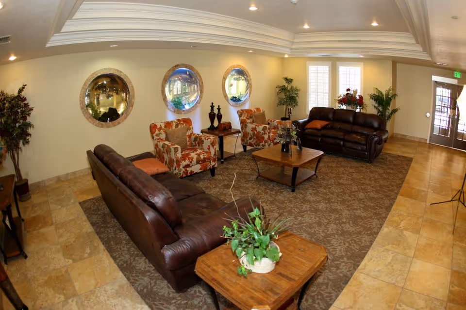 A cozy living room area with two brown leather sofas and two floral-patterned armchairs arranged around a wooden coffee table on a patterned rug. The walls are light-colored with three round mirrors, and there are several potted plants and floral arrangements adding greenery to the space. The floor is tiled, and there is a door with glass panels letting in natural light.