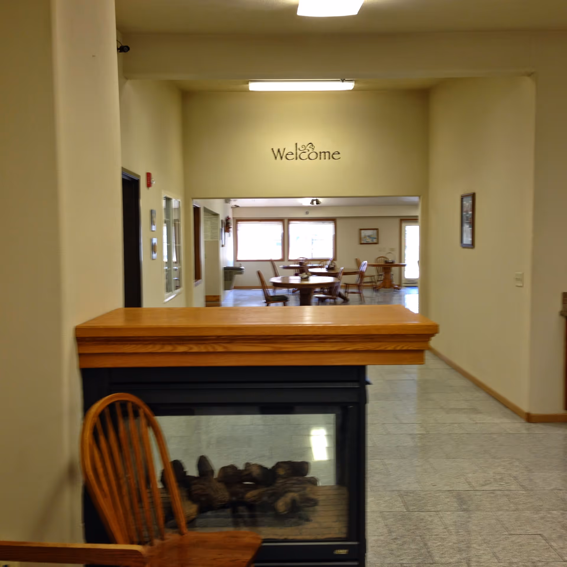 Interior view of a senior living facility entry with a wooden counter and fireplace in the foreground and a dining area with tables and a 'Welcome' sign in the background.