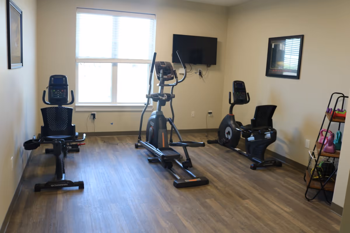 A small fitness room with three exercise machines including two recumbent bikes and an elliptical trainer. There is a large window with blinds letting in natural light, a wall-mounted TV, framed pictures on the walls, and a shelving unit holding colorful kettlebells and other small exercise equipment.