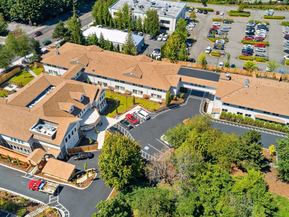Aerial view of Vineyard Park of Puyallup senior living facility showing multiple connected buildings with tan roofs, surrounding parking lots with cars, landscaped greenery, and trees.