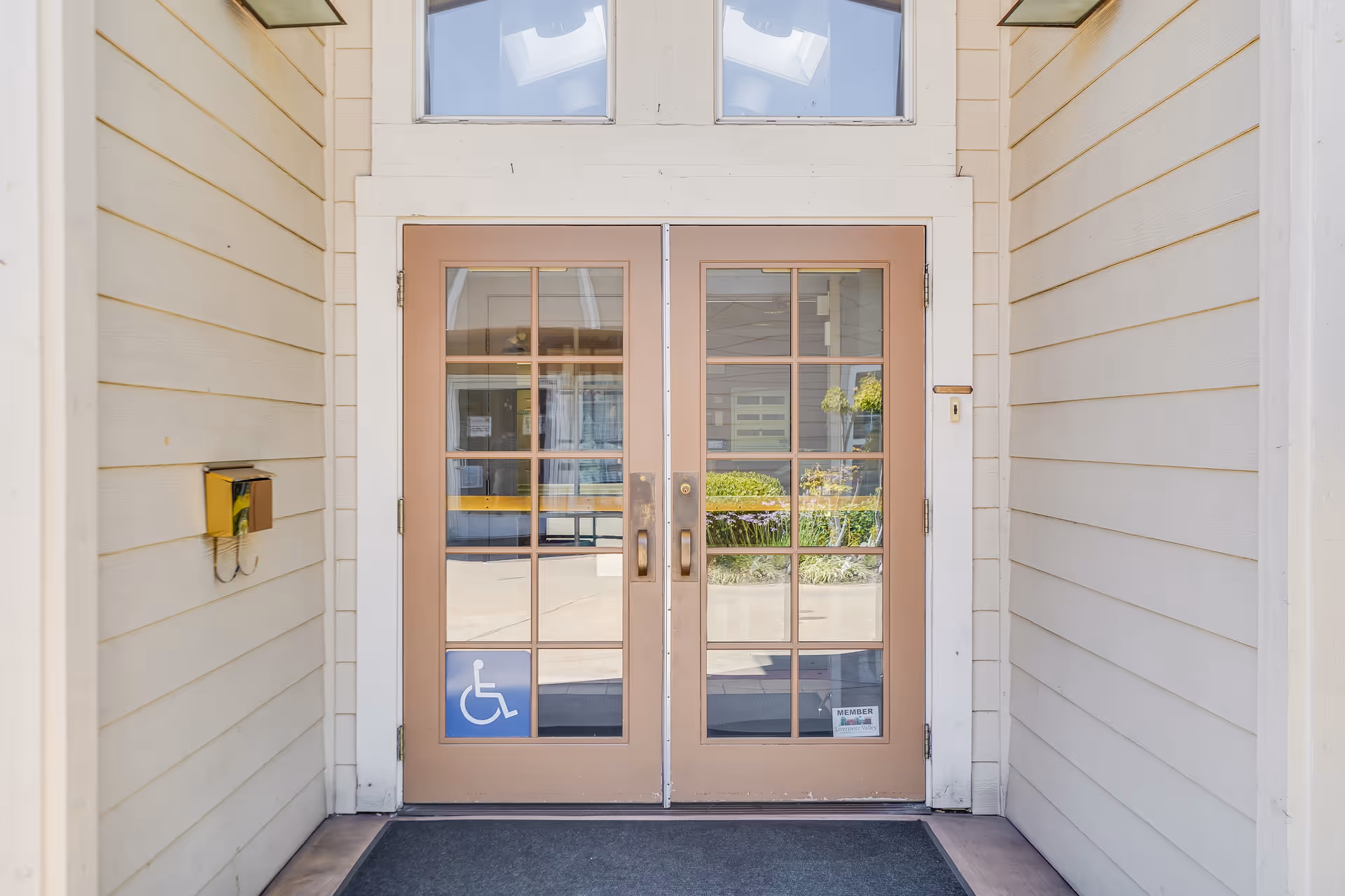 Double glass doors with brown frames at the entrance of a building, featuring a wheelchair accessibility sign on the left door and a membership sticker on the right door. The doors are set within a beige siding exterior with a black doormat in front.
