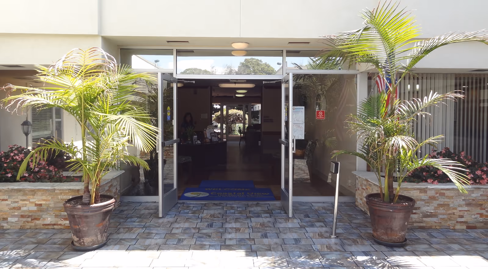 Entrance to Coastal View Healthcare Center with double glass doors open, flanked by two large potted palm plants. The exterior features stone wall accents and a tiled walkway leading inside. Inside, a reception desk is visible with a person standing behind it.