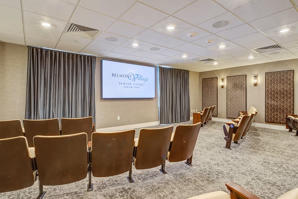 Interior view of a senior living facility's small theater or presentation room with rows of brown cushioned chairs facing a large screen displaying the Belmont Village Senior Living Hunters Creek logo. The room has beige walls, patterned wall panels with sconces, and gray curtains covering windows.
