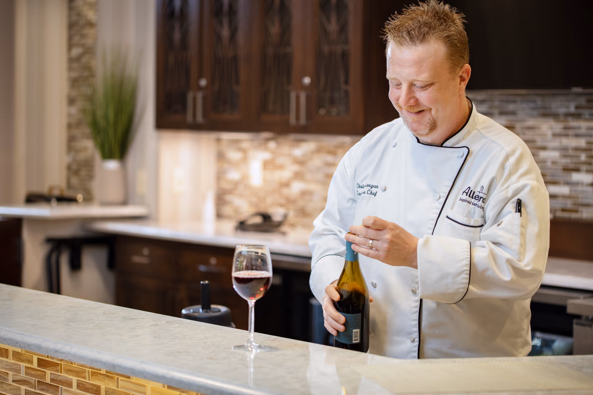 A chef in a white uniform with the Allegro logo is opening a bottle of wine behind a kitchen counter with a glass of red wine placed on the counter. The kitchen has dark wooden cabinets and a tiled backsplash.