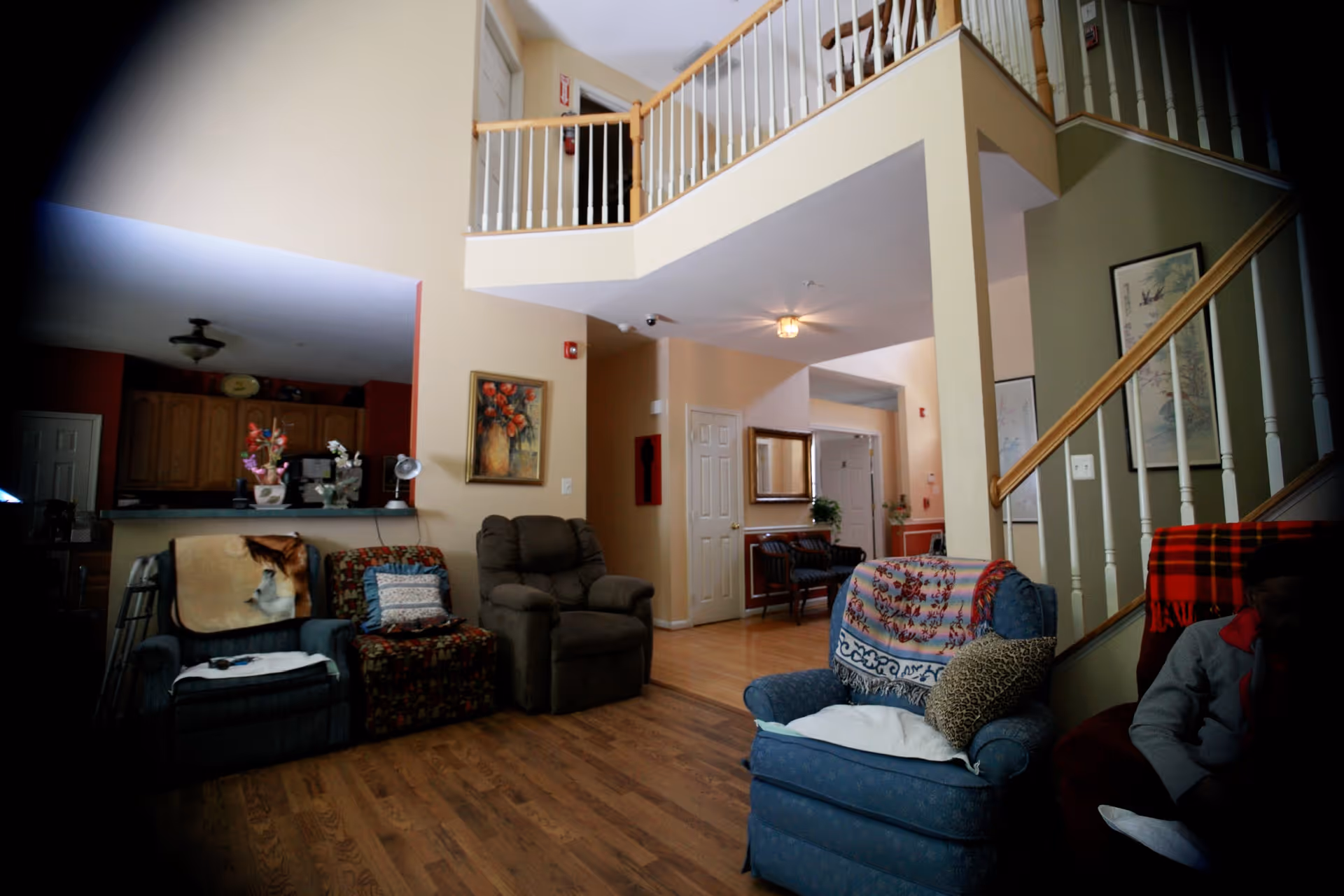 Interior view of a senior living facility common area with multiple armchairs, a staircase with white railings, wooden flooring, and a kitchen area in the background. The space has beige walls, framed artwork, and a second-floor balcony overlooking the room.