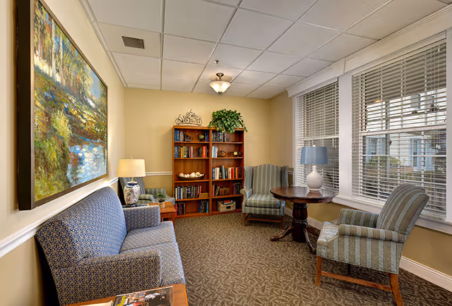 A cozy sitting area in a memory care community featuring a blue patterned sofa, two striped armchairs, a round wooden table with a white lamp, a wooden bookshelf filled with books and decorative items, a large painting of a nature scene on the wall, and windows with white blinds letting in natural light.