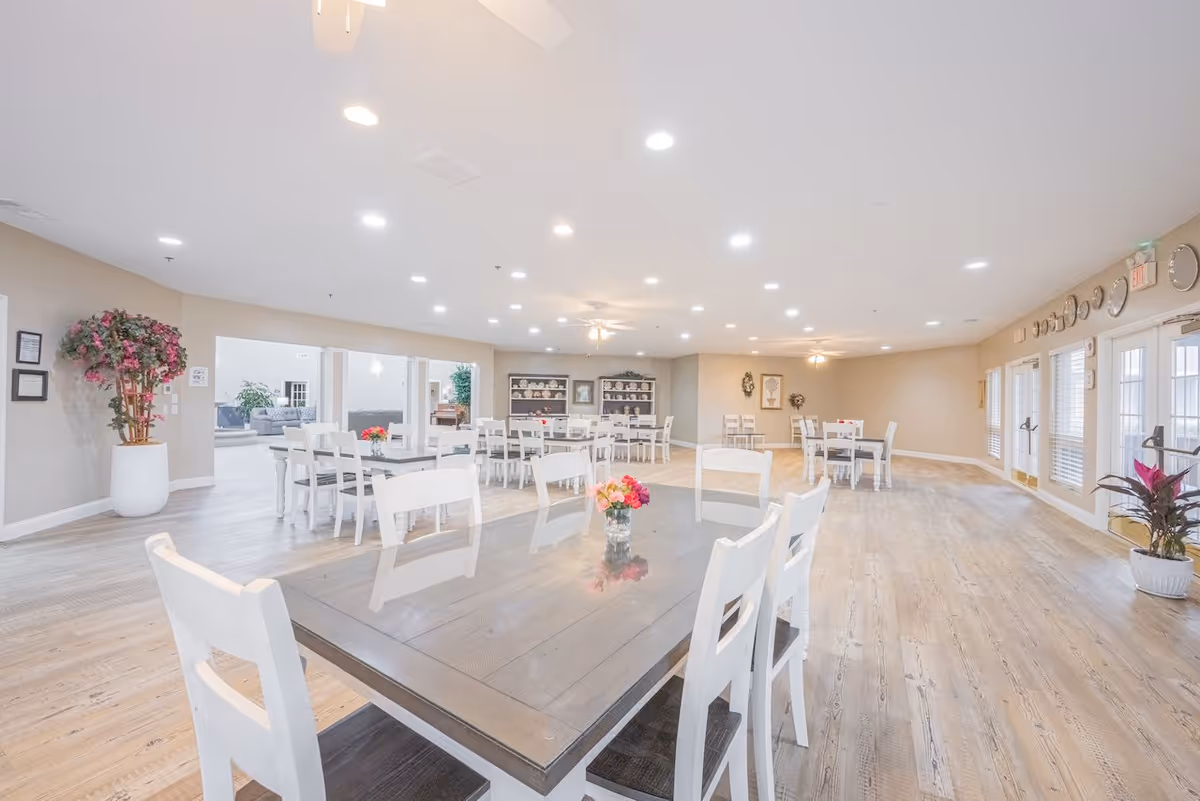 A spacious and bright dining room with multiple tables and white chairs arranged neatly. The room features wooden flooring, ceiling fans, recessed lighting, and large windows with blinds. There are decorative plants and flowers on the tables and around the room, along with wall clocks and framed pictures on the walls.