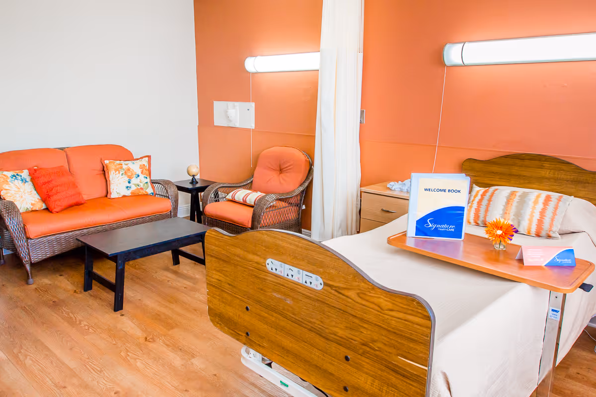 A senior living bedroom with a hospital-style bed and overbed table, and an orange seating area with a sofa and chair.