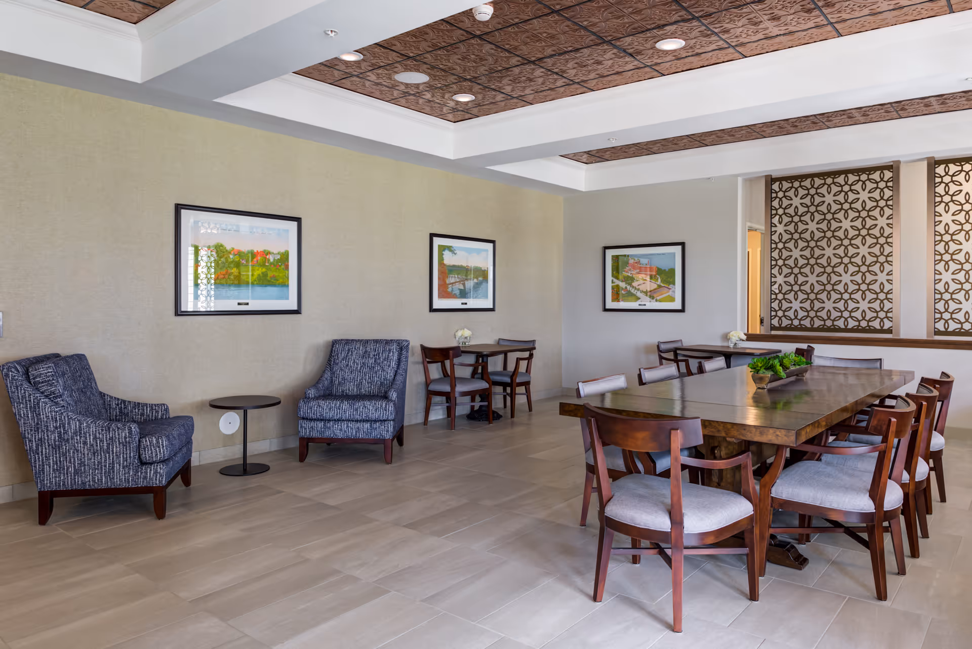 A spacious, well-lit common area in a senior living facility featuring a large wooden table surrounded by chairs, two upholstered armchairs with a small round side table between them, and two smaller tables with chairs against the wall. The walls are decorated with framed artwork, and the ceiling has a decorative wooden panel design.