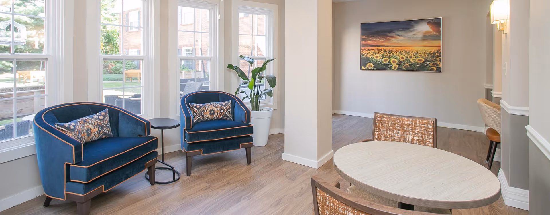 A bright and cozy sitting area with two blue armchairs featuring patterned cushions, a small round black side table between them, a large potted plant near tall windows showing an outdoor garden, and a round wooden table with chairs in the foreground. A colorful sunflower field painting hangs on the wall.