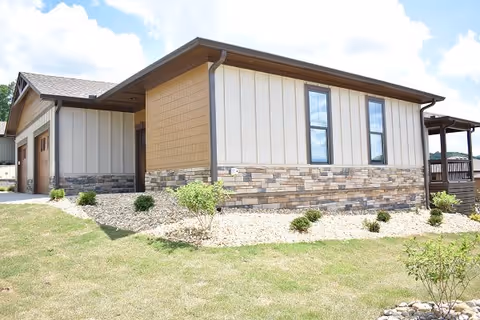 Exterior view of a single-story building with stone veneer, vertical siding, a garage, and a small landscaped yard under a blue sky.