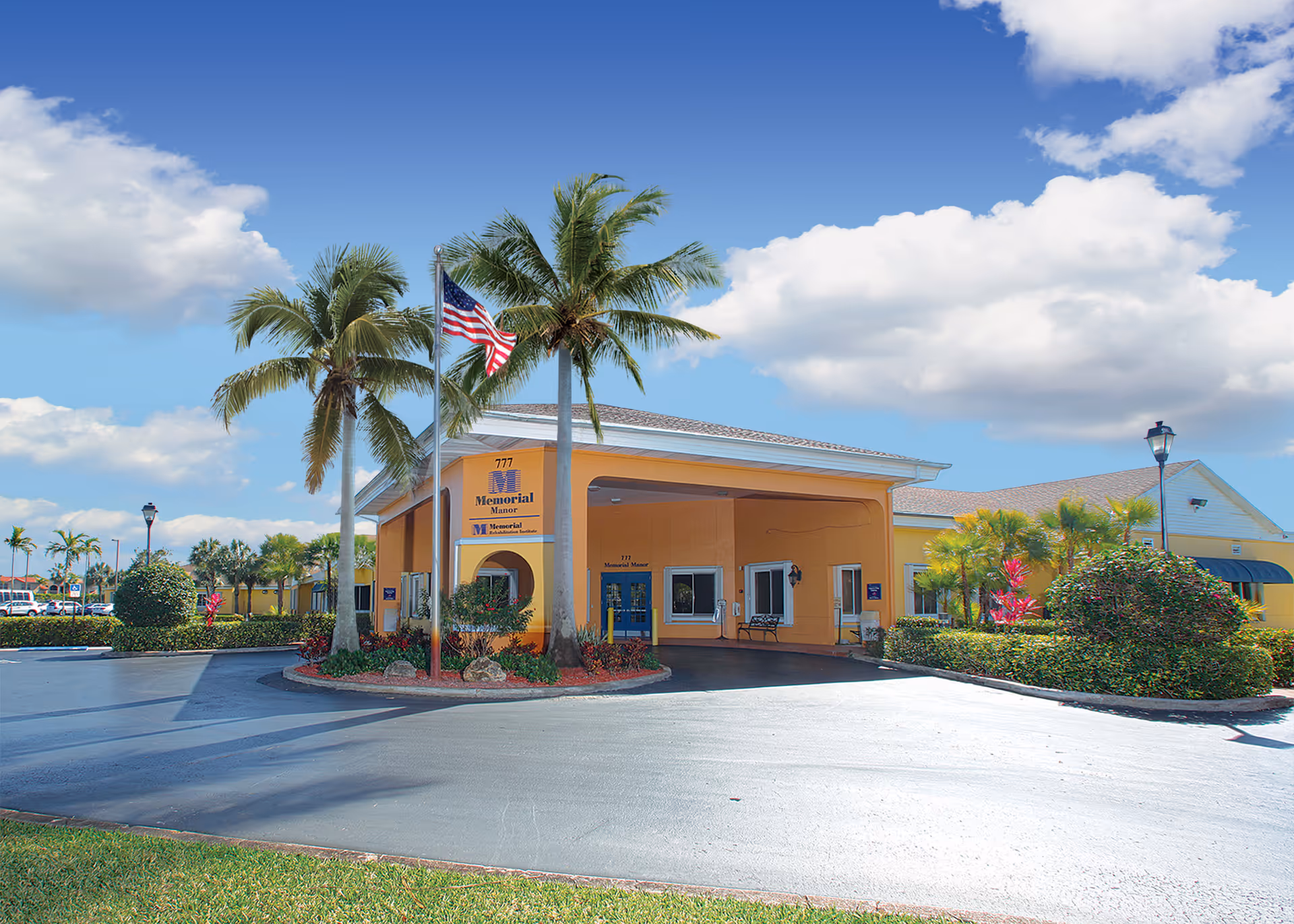 Exterior view of Memorial Manor senior living facility with a yellow building, palm trees, an American flag, and a clear blue sky with some clouds.