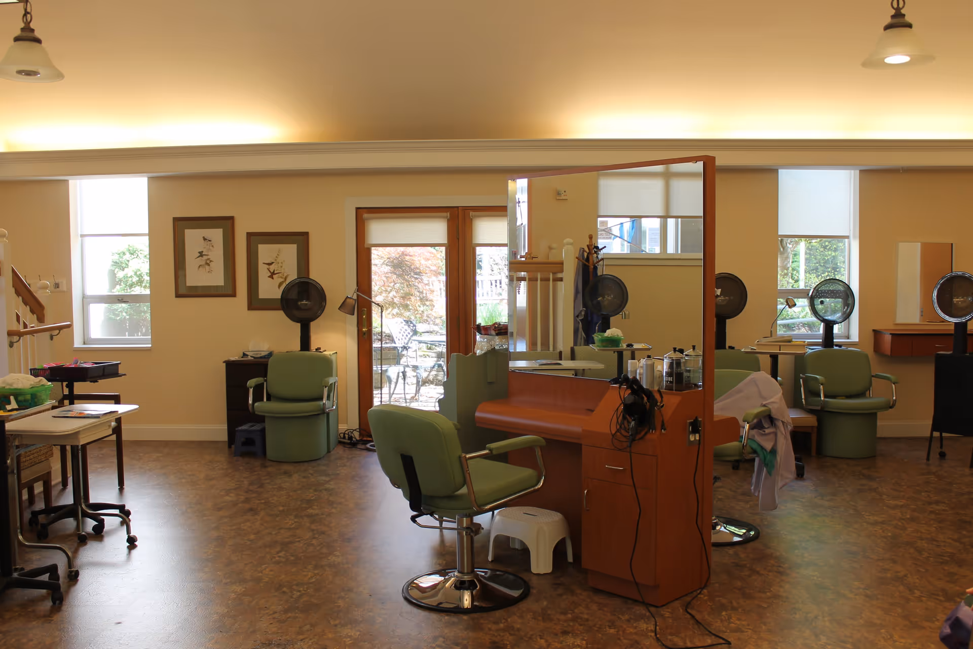 Interior of a salon area in a senior living facility with green salon chairs, hair dryers, a large mirror, and a door leading to an outdoor patio. The room has warm lighting, framed bird artwork on the wall, and windows letting in natural light.