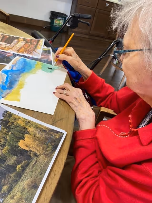 An elderly woman wearing glasses and a red sweater is seated at a wooden table, painting a landscape on a sheet of paper. She is holding a paintbrush and working on a colorful scene with blue, yellow, and green hues. There are printed photos of nature scenes on the table near her. A walker is visible in the background.