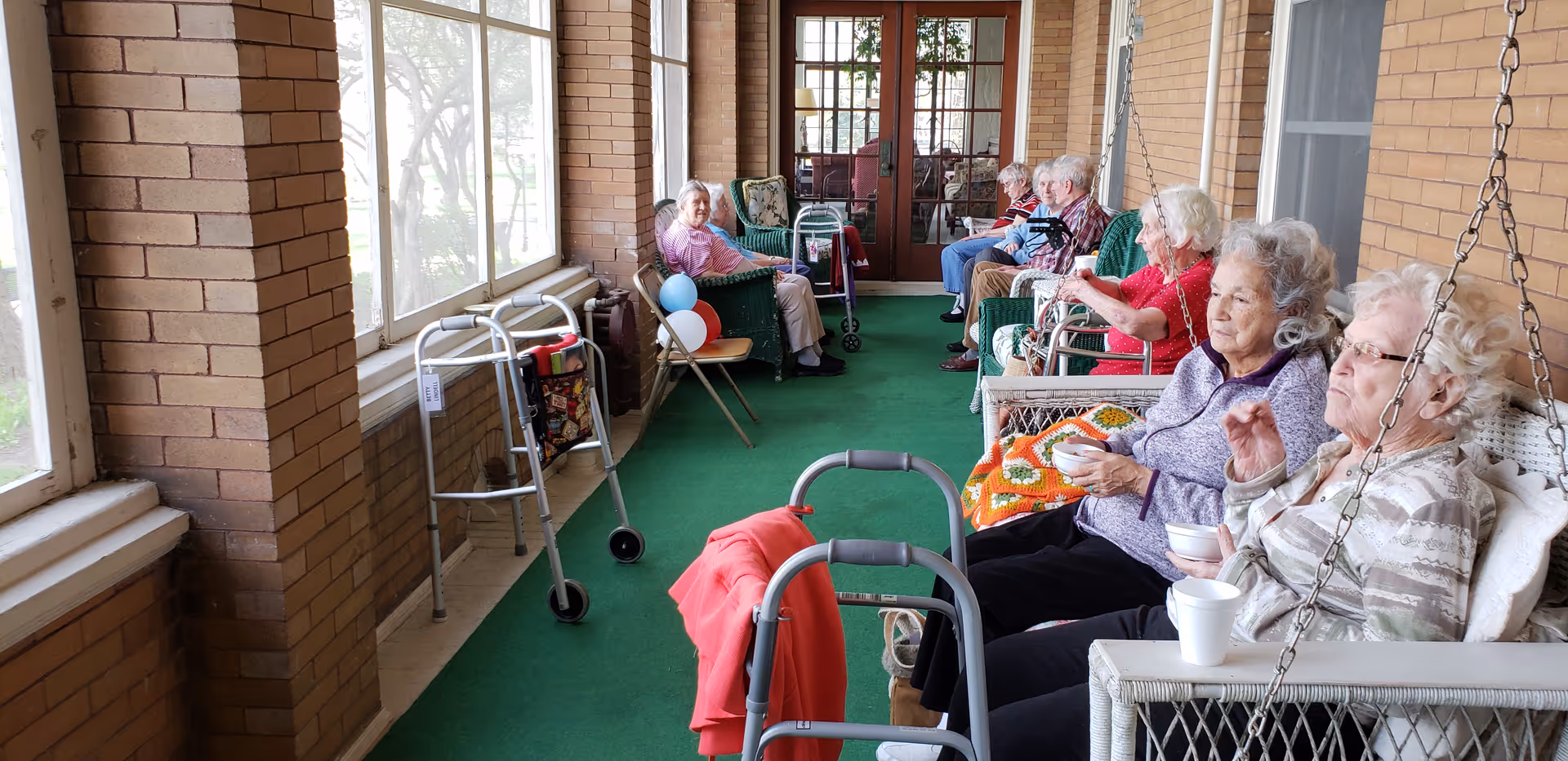 A group of elderly people sitting on wicker chairs and swings on a covered porch with green carpet. Several walkers are positioned nearby, and some individuals are holding cups and bowls. The porch has large windows on one side and brick walls with glass doors at the far end.